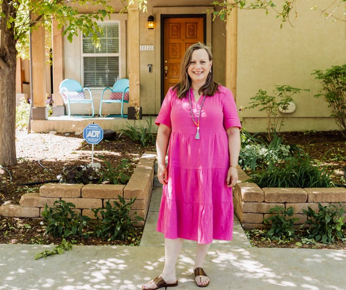Person standing in front of a house with a garden, wearing a pink dress smiling at the camera. 