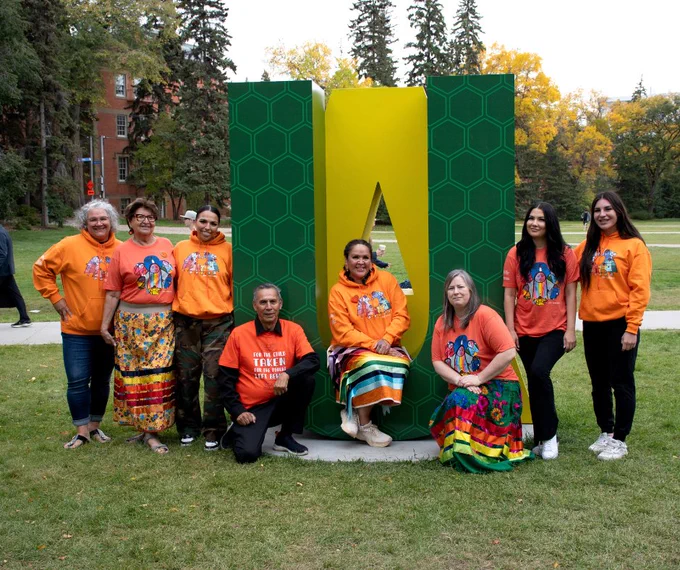 Group of eight people from First People's House standing in front of a large green and gold 'UA' sculpture on University of Alberta North Campus, wearing orange shirts.
