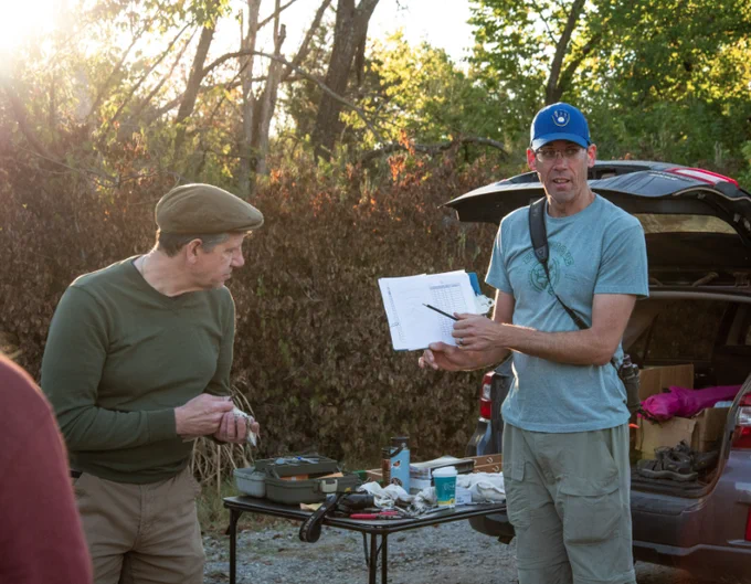 Two white guys standing in front of a card table that is crowded with bird banding supplies. One guy is holding a bird and the other is pointing to a datasheet on which measurements from the bird will be entered. Judging from their hats, the guy with the bird has been to Ireland at some point, and the tall guy is a Milwaukee Brewers fan. Photo by Clay Billman. 