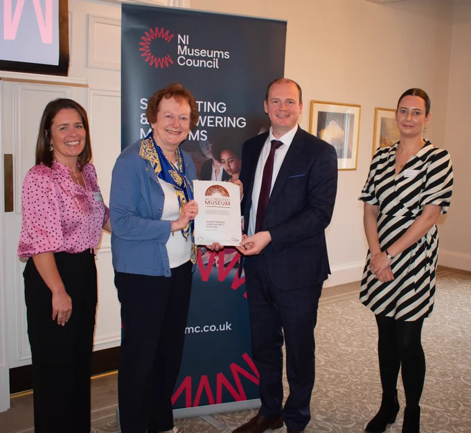 Four smiling people standing in front of an NI Museums Council banner, the two people in the center holding a museum accreditation certificate on either side.