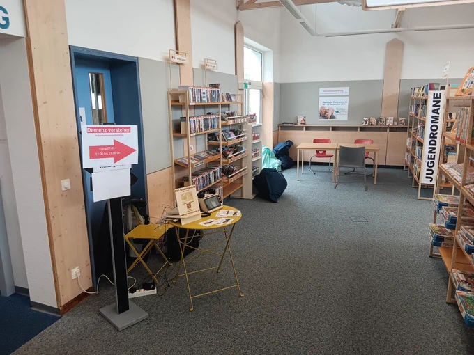 Der Blick in den linken Teil der Neu-Ulmer Stadtbibliothek. Am Ende des Gangs zwei Tische mit Stühlen und einem Plakat an der Wand, das auf den Informationsnachmittag der Seniorenberatung zum Thema Demenz hinweist. Links im Gang steht ein Poller mit einem Wegweiser-Pfeil. Direkt daneben ein kleiner gelber runder Tisch mit einem der Grannophone-Prototypen und einem lasergravierten Infoschild zum Projekt.