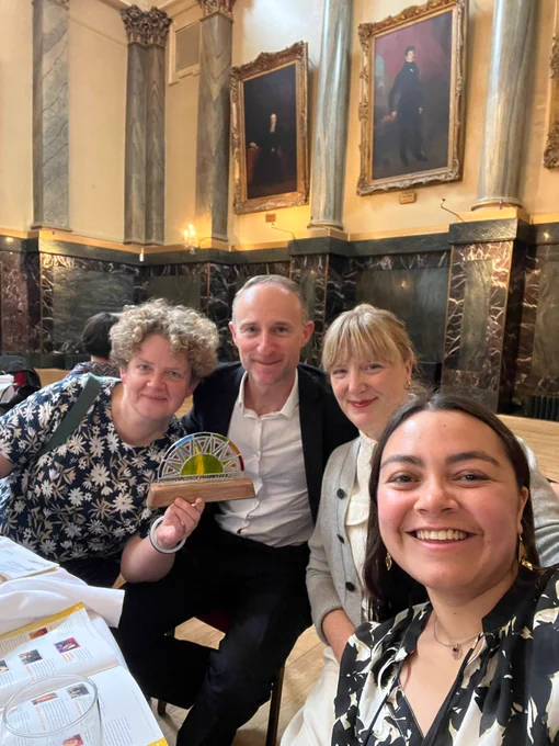 Four people are seated at a dinner table, smiling and holding a colourful, semi circle award, The award is made from yellow and orange stained glass and is set into a wooden base. In the background are some large portrait paintings.