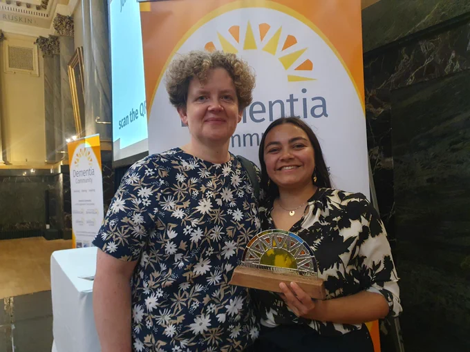 Two people stand holding a colourful award in from of an orange and yellow banner that reads 'Dementia Community'. 