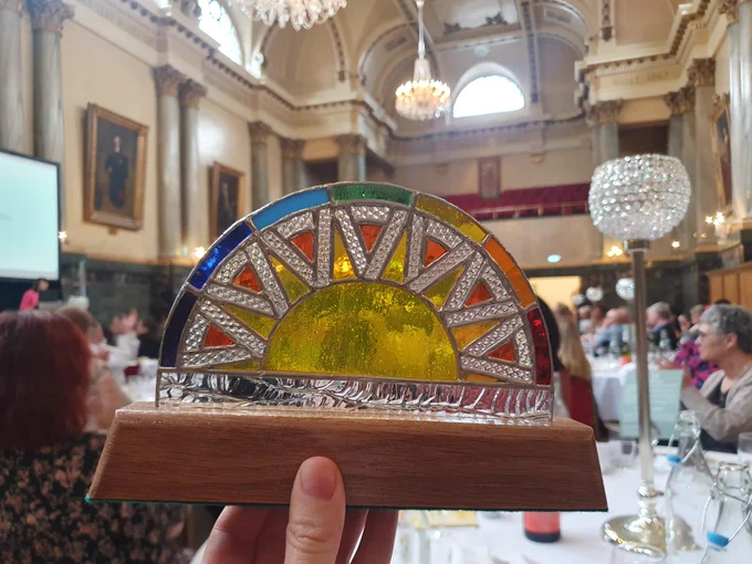 A close up photo of a colourful, semi circle award. The award is made from yellow and orange stained glass and is set into a wooden base. In the background are people sat at dinner tables in a large hall.