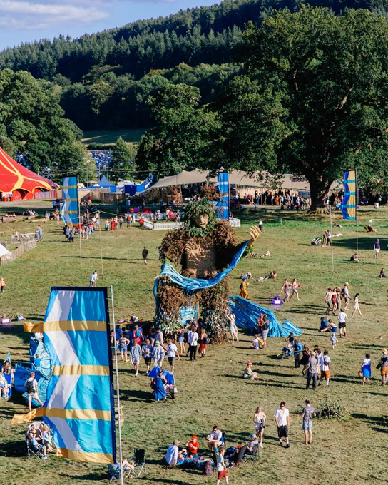 Outdoor festival scene with a large Green Man sculpture and people walking around.