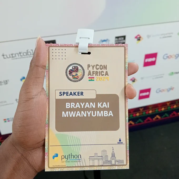 A speaker Badge for PyCon Africa 2024 with the name "Brayan Kai Mwanyumba" on it. In the background is a blurred backdrop banner with logos of sponsors of the conference.