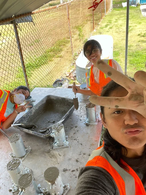 Students mix concrete to form small cylinders in an outdoor shaded area near a fence