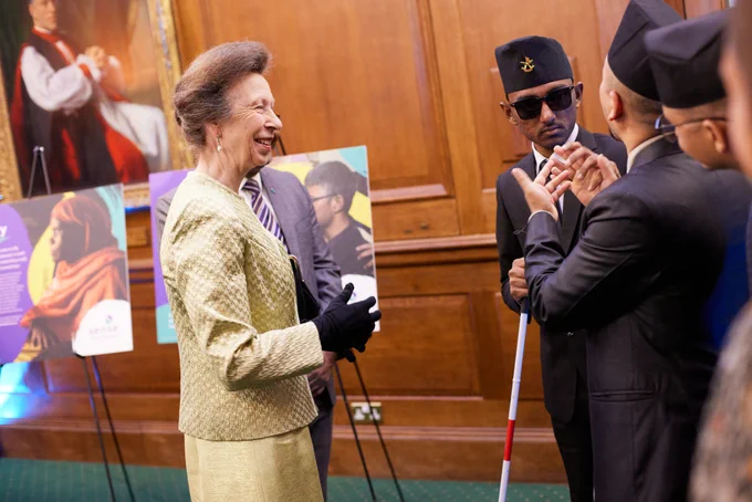 HRH Princess Anne interacts with young man with deafblindness at an event, both smiling and engaging in conversation. There are posters and attendees in the background.