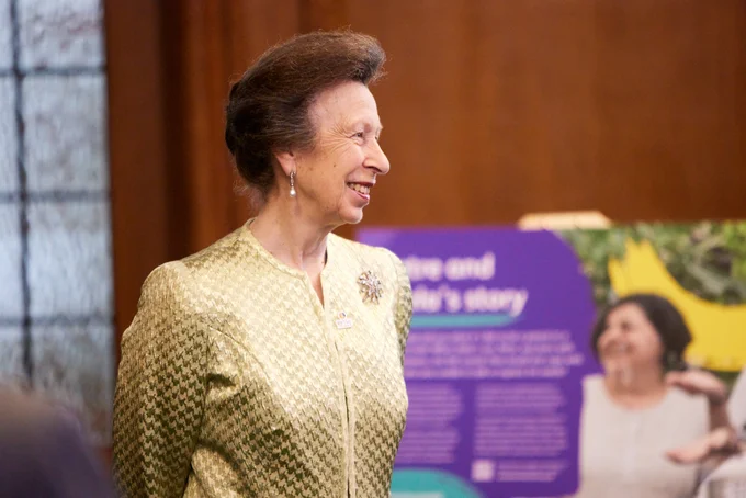 HRH Princess Anne smiling during an event, wearing a gold jacket, with a poster in the background.