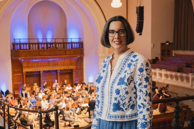 Sarah Bardwell, a woman with brown hair and glasses and wearing a white cardigan with a blue floral pattern, stands on the Gallery at Cadogan Hall with the Orchestra rehearsing below her.