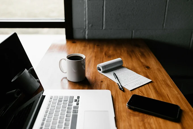 Desk with laptop, coffee, notepad and phone. Image by Andrew Neel on Unsplash.