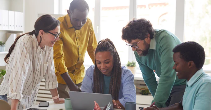 Diverse group of people gathered around a laptop, smiling & interested
