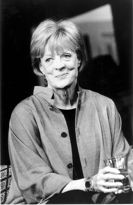 A black and white image of a person sitting down with short hair, smiling off-camera. They are wearing a grey shirt open with a black t-shirt underneath, holding a glass of water. 