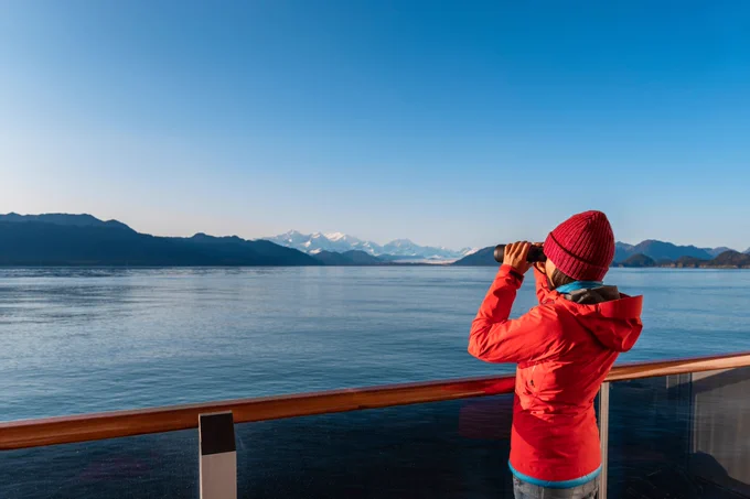 A woman in winter attire (jacket and hat) using a pair of binoculars looks off into the distance at some mountains. She is standing on what appears to be the deck of a boat.