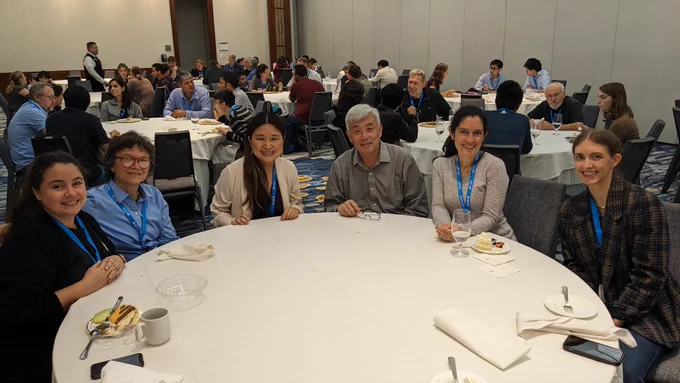 Six attendees of ASBMB's conference on transcriptional regulation sit at a round table in a conference room. There are five other tables behind them at which attendees are seated eating lunch and conversing. 