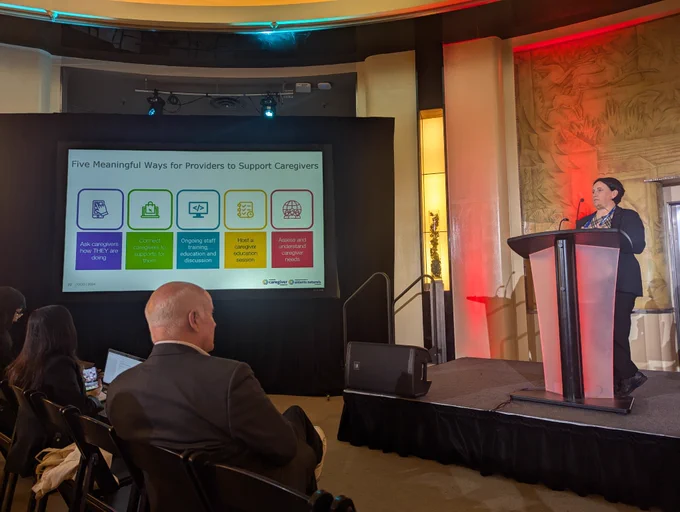 Adrien McKenna speaking on stage. On the left is a slide talking about Five Meaningful Ways for Providers to Support Caregivers. 