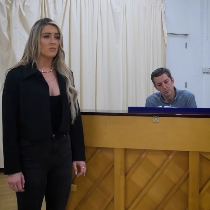 A photo showing a performer and a pianist in a rehearsal room, stood and sat around a piano 