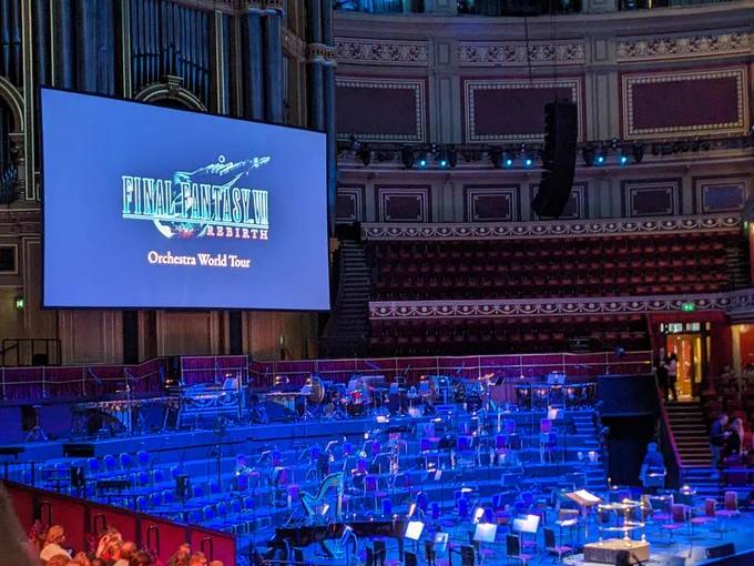 A photograph of an orchestra setup. The screen behind the orchestra says "Final Fantasy Rebirth Orchestra World Tour"