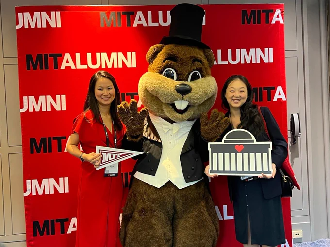 Two MIT alumni stand with Tim the Beaver mascot in front of a red MIT Alumni backdrop. One holds a pennant and the other holds a mini replica of the MIT dome.
