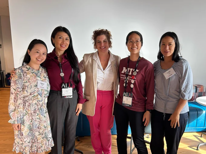 Five alumni smiling at ALC, wearing name tags, standing in a room with a bright, modern decor.