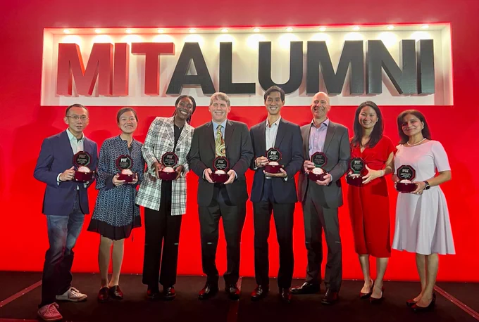 Group of seven alumni holding awards, smiling, in front of a red 'MIT ALUMNI' sign, at an event.