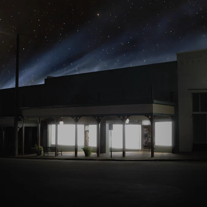 Nighttime street view of a small-town commercial building with bright, illuminated storefront windows. The sky above features a backdrop of stars, adding to the serene and quiet atmosphere. The building has a vintage design with wooden columns and a covered walkway, hinting at historical charm. The bright interior lights of the shop contrast sharply with the dark street, making the space feel welcoming and lively even in the late hours.