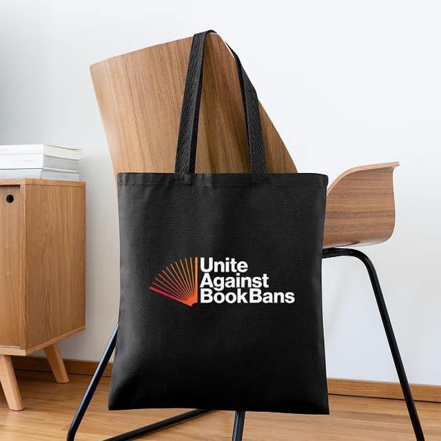 A black Unite Against Book Bans tote is slung over a wooden chair.