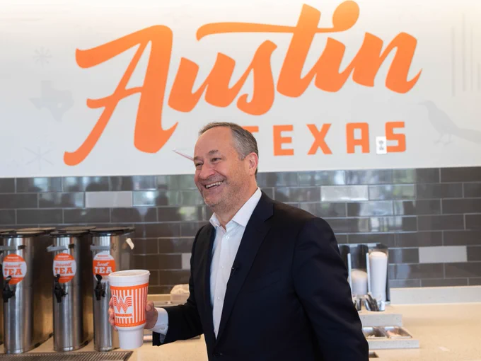 Second Gentleman Doug Emhoff smiling in a Whataburger holding a cup, standing in front of a counter with beverage dispensers. In the background is a large sign that reads "Austin Texas.
