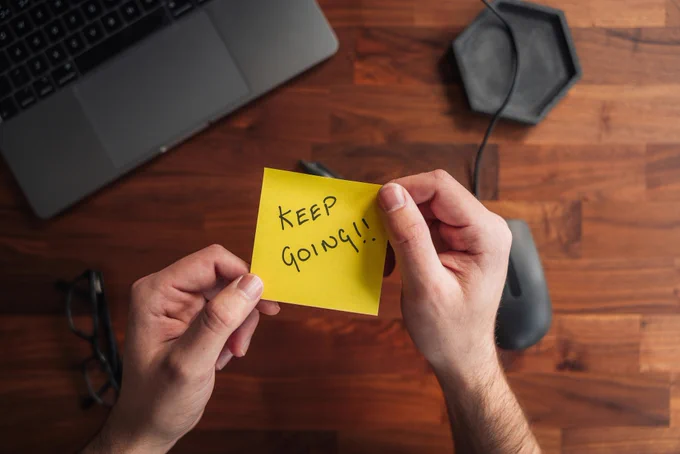 A person holding a yellow PostIt note with both hands. On the note, written in black ink is the next "Keep Going!!"