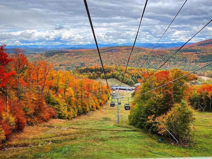 View from a ski lift overlooking vibrant fall foliage at Gore Mountain, with cable lines and ski lift chairs in the foreground, leading down to a small base area.