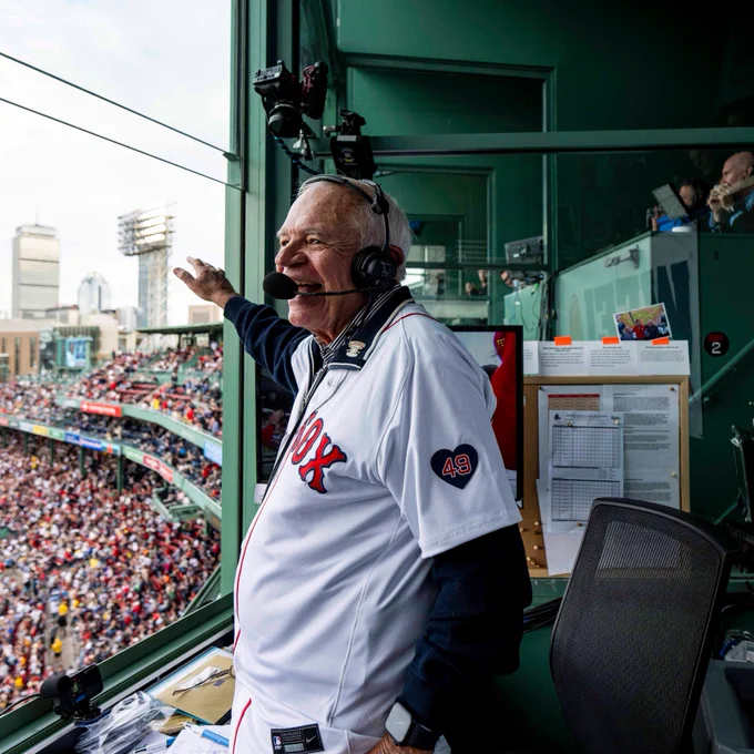Joe Castiglione waves to the crowd from the radio booth at Fenway Park. He wears a radio headset and a white home Red Sox jersey.