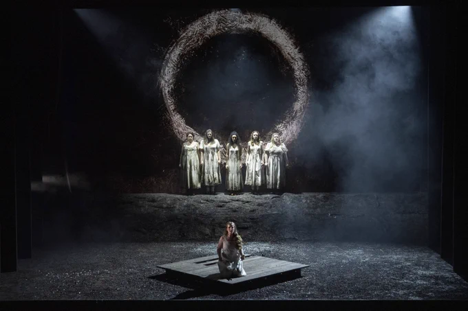 A person kneels on a dimly lit stage with six figures in white robes standing in the background in front of a large circular structure, shrouded in smoke.
