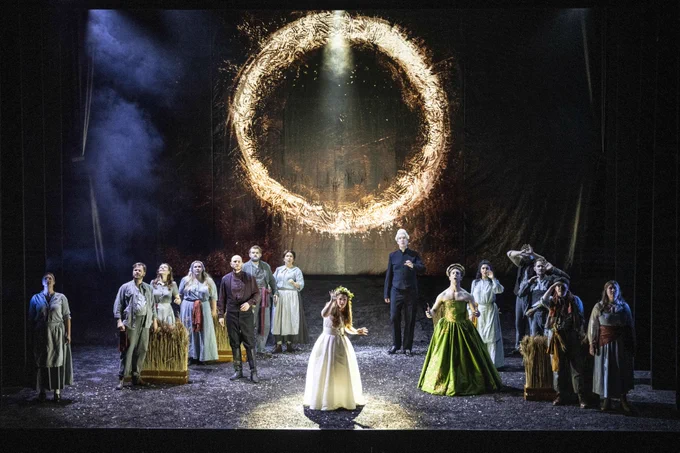 A theatrical stage scene with performers in historical costumes, standing and interacting under a circular, fiery light display. The backdrop features dramatic lighting and fog effects.