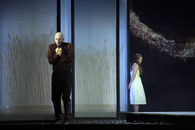 A man wearing black is standing at the front of the stage while a women wearing a white dress is standing far behind him leaning against a wall