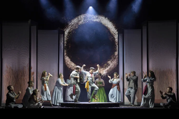 The English Touring Opera company poses together, showcasing their vibrant costumes and joyful expressions on stage with a ring of fire behind them.