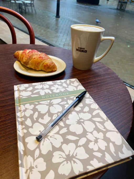 Photo shows a grey floral notebook on a dark wooden table, next to a mug of coffee and a croissant. There is a view of the rainy street outside the window. 