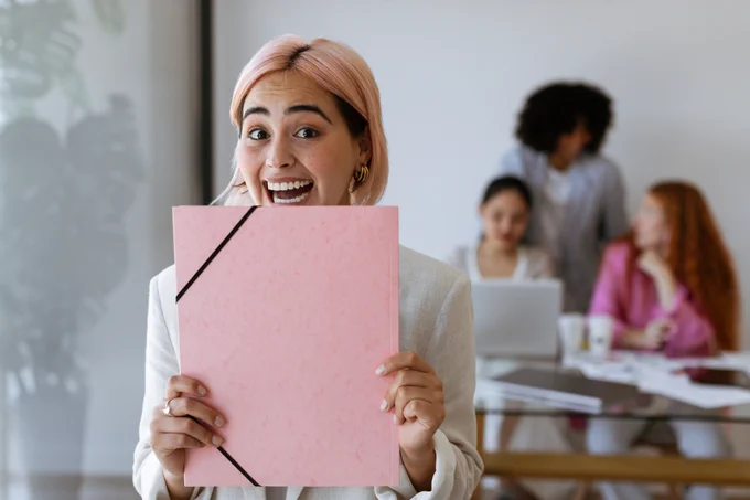 Image of a smiling young woman in an office holding a folder up to her face as if celebrating a success at work.