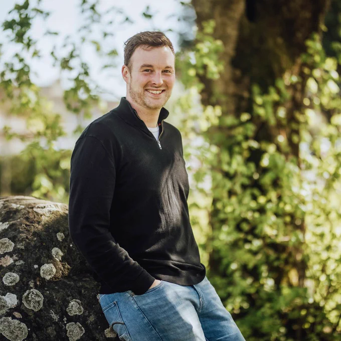 A male MIC mature student leaning against a large stone. He is smiling and has his hands in the pockets of his blue denim jeans. He has short brown hair and is wearing a black zip neck jumper and white t-shirt. The sun is shining through the leaves of the trees in the background. 