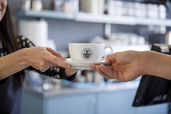 A coffee cup, with the Explore Cafes logo, is being passed between two people