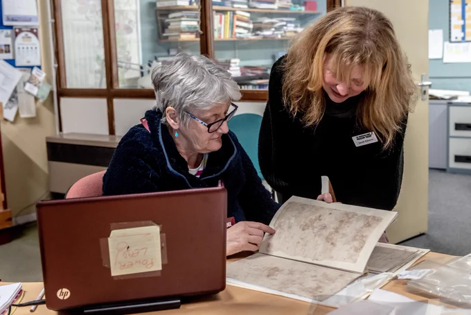 Photo of two women reading records in an archive. One woman is sitting and one is standing looking over the documents.