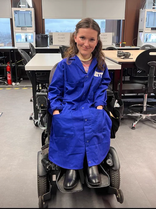 full body image of Anna, a young white woman in her black electric wheelchair, wearing the new, adapted lab coat that fits her petite frame well and doesn’t get in the way of her lab-work. The coat is a vibrant royal blue.