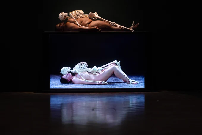 Gallery view of Madinah Farhannah Thompson re-performing 'Nude with Skeleton’ at the Royal Academy of Arts. Madinah is lying on the floor naked, with a human skeleton placed on top of her.