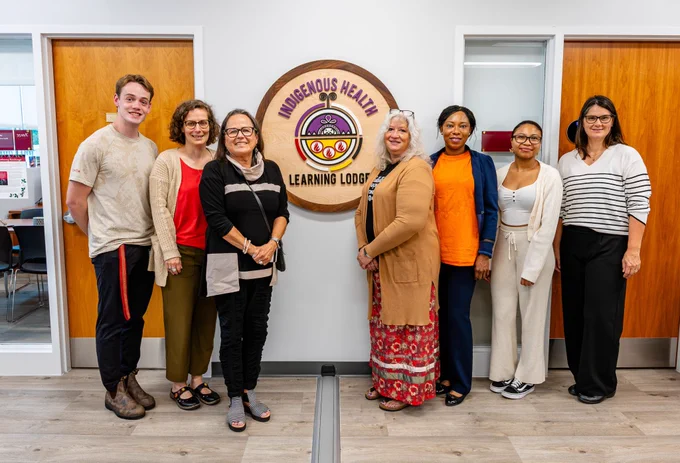A group of seven McMaster community members standing on either side of signage that reads, ‘Indigenous Health Learning Lodge.’ 