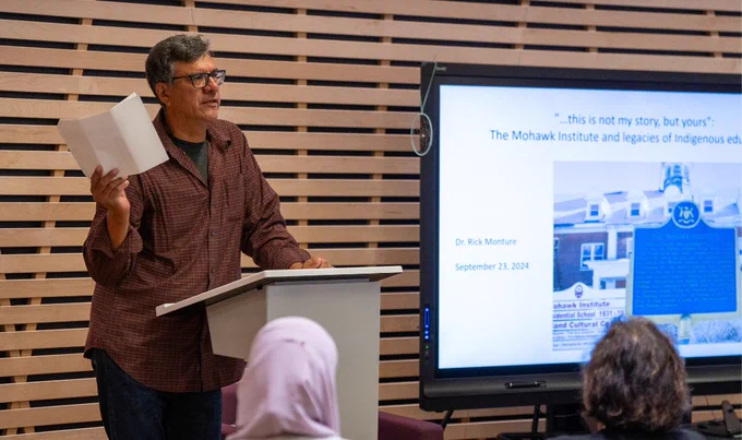 McMaster Professor Rick Monture speaks at a podium. To his left is a screen showing a presentation that includes an archival image of the Mohawk Institute Residential School. 
