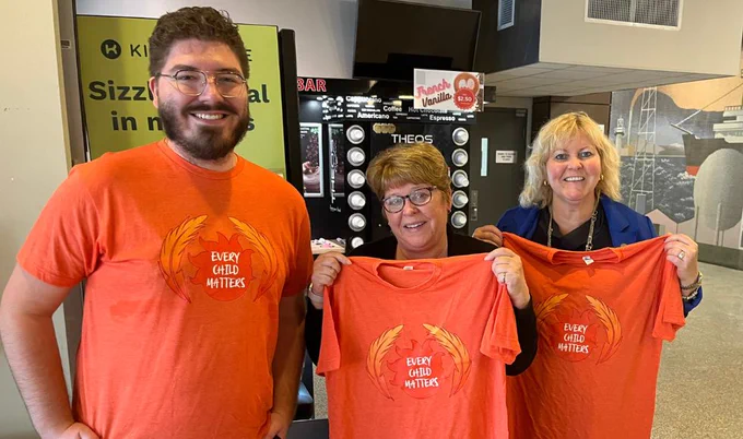 Three people, either wearing or holding up an orange shirt, smiling at the camera 