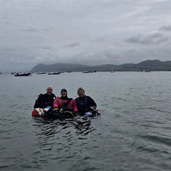 Three scuba divers in wetsuits floating on the surface of the water with hills and cloudy skies in the background.