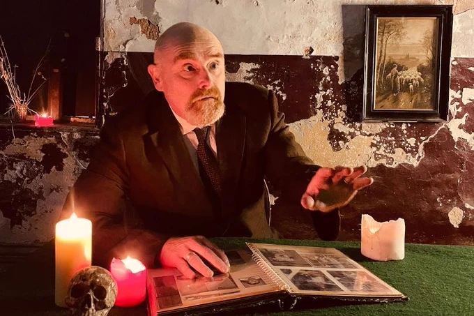 Photo of Vic Llewellyn sitting at a table with an open photo album, three large lit candles and a skull on it. The wall behind him is beaten up and scuffed, with a framed drawing of a rural scene hung on it. Vic is wearing a black suit and tie and is holding one hand up in front of him as if in the middle of telling a story.