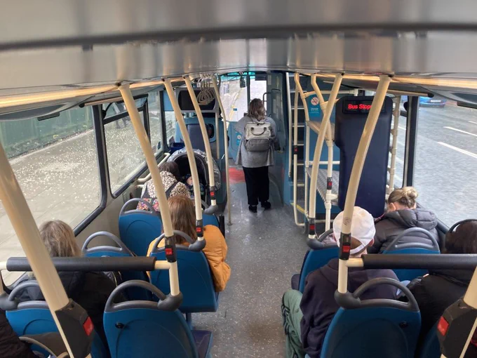 Interior of a bus with passengers seated and one person standing near the exit door; view from the rear of the bus towards the front.