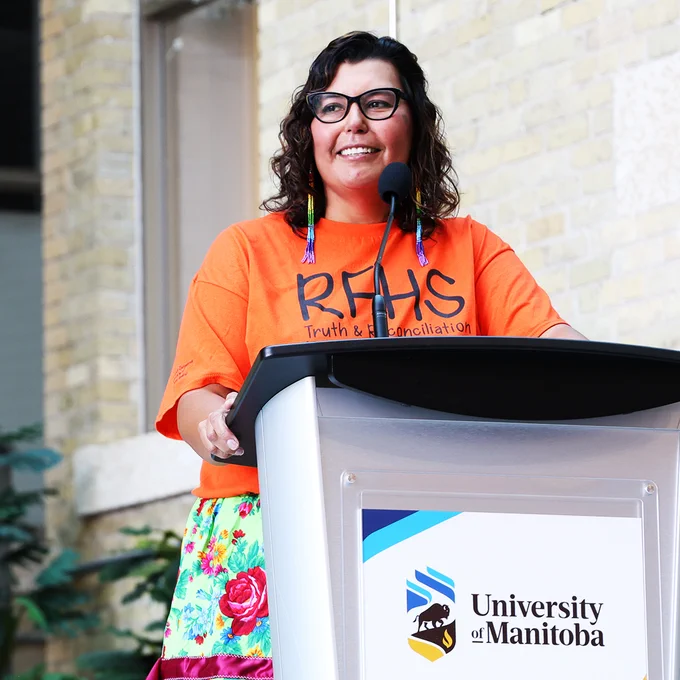 Photo of Dr. Marcia Anderson, smiling, wearing an orange shirt reading RHFS and a colourful ribbon skirt. She is standing behind a podium with the University of Manitoba logo.