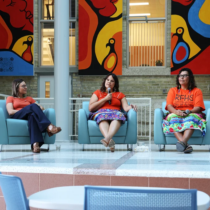Picture of a panel of three women, seated in front of a colourful indigenous mural left to right, Dr. Vanessa Poliqiun, Dr. LIsa Monkman and Dr. Marcia Anderson.  All three are wearing orange shirts, Drs Anderson and Monkman are also wearing ribbon skirts.